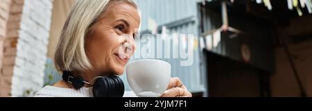 Une femme aux cheveux gris aime le café et sourit tout en portant des écouteurs dans un espace confortable. Banque D'Images