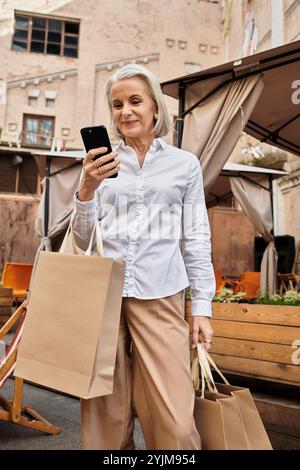 Une femme sophistiquée vérifie son téléphone tout en transportant des sacs à provisions à l'extérieur dans un endroit charmant. Banque D'Images