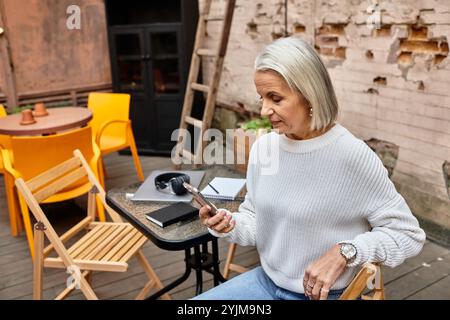 Dans un cadre extérieur charmant, une femme aux cheveux gris se détend tout en utilisant son smartphone. Banque D'Images