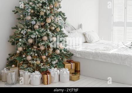 Matin de vacances dans une chambre lumineuse et décorée avec arbre de Noël avec des ornements et des lumières et des boîtes cadeaux emballées rassemblées par le lit avec literie blanche, créa Banque D'Images