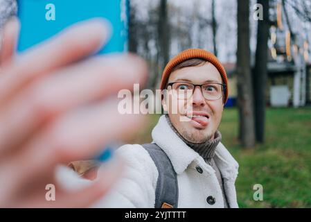 Jeune homme touriste dans un parc de la ville prenant un selfie drôle, sortant sa langue. Parfait pour le partage sur les réseaux sociaux pour se souvenir d'une aventure amusante. Moment urbain joyeux capturé sur smartphone Banque D'Images