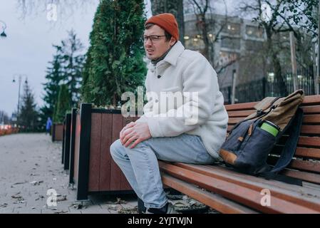 Un jeune homme portant une veste blanche et un bonnet orange est assis sur un banc de parc, éprouvant de l'inconfort et tenant son genou dans la douleur, peut-être en raison d'une blessure ou de l'arthrite Banque D'Images