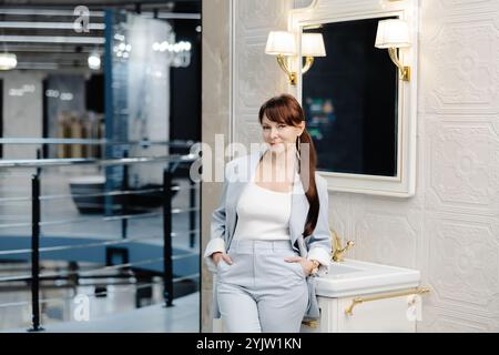 Une femme en costume bleu ajuste le robinet d'un évier blanc dans une salle d'exposition de salle de bains. La salle de bains présente des murs en carrelage blanc et un grand miroir décoré Banque D'Images