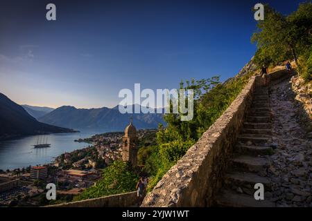 Randonnée sur le sentier menant aux ruines de la forteresse de Kotor, avec les gens dans l'escalier, la tour de l'église en vue, et la ville s'étendant en dessous par une journée d'été ensoleillée Banque D'Images