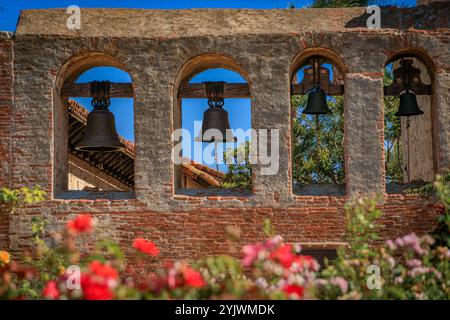San Juan Capistrano, États-Unis - 6 août 2023 : trois cloches historiques accrochées dans des fenêtres cintrées, jardin fleuri au premier plan, Mission San Juan Capistrano Banque D'Images