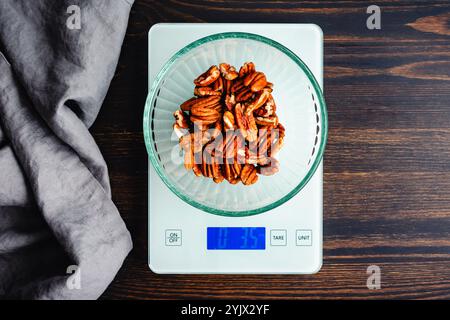 Peser les moitiés de noix de pécan sur une balance de cuisine numérique : vue de dessus des noix dans un bol en verre pesé sur une balance alimentaire sur une table en bois Banque D'Images