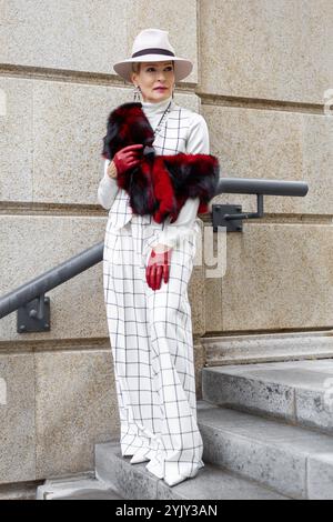 Femme mature à la mode marchant à l'extérieur dans la ville habillée d'élégant costume blanc à carreaux et pantalon avec chapeau Fedora, accessoires de fausse fourrure mise en valeur Banque D'Images