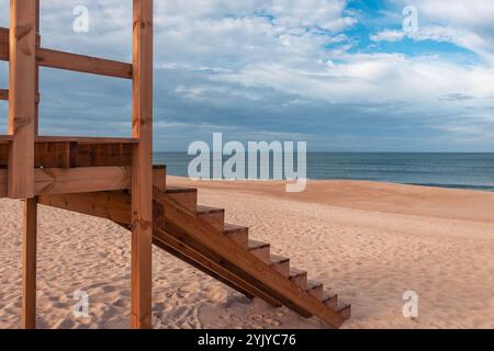 Gros plan d'escaliers de tour de sauveteur en bois sur une plage de sable sereine au Portugal, avec l'océan Atlantique en arrière-plan sous un ciel bleu partiellement nuageux Banque D'Images