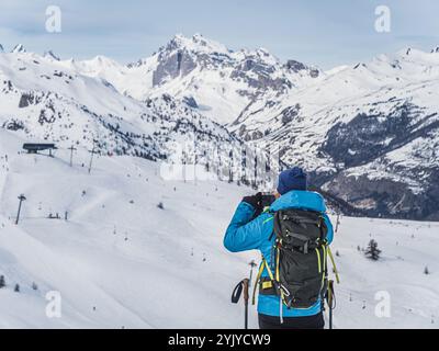 Un randonneur seul s'arrête pour capturer les superbes paysages de montagne enneigés sur un smartphone, entouré d'arbres enneigés et de pics accidentés sur un sentier d'hiver Banque D'Images