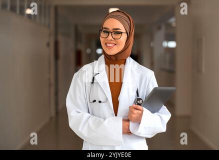 Portrait de femme musulmane heureuse médecin ou infirmière portant un manteau blanc avec stéthoscope dans le couloir de l'hôpital tenant la tablette numérique regardant la caméra et Banque D'Images