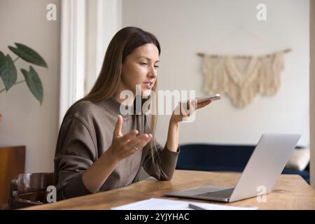 Femme d'affaires assise à table avec ordinateur portable, à l'aide d'un smartphone, enregistrement de la messagerie vocale Banque D'Images