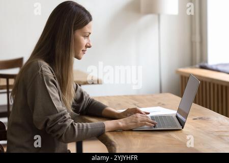 Vue de côté femme concentrée assise à table, travaillant sur ordinateur portable Banque D'Images