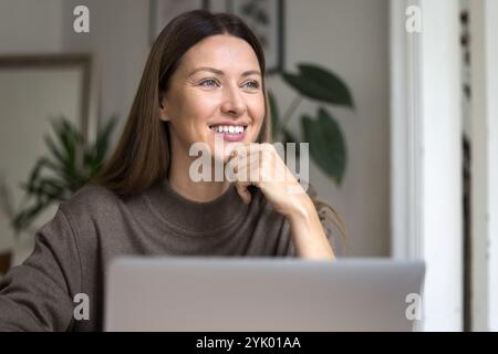 Femme assise devant l'ordinateur portable, regardant loin avec le sourire Banque D'Images