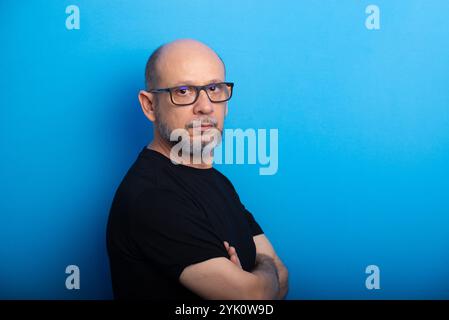 Barbu, homme chauve portant des lunettes de vue avec les bras croisés posant pour la photo sur fond bleu. Prise de vue en studio. Banque D'Images