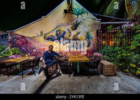 Le directeur général du MosaCaña Bar & Kitchen est assis dans l'espace extérieur, Penstraat 41, Willemstad, devant la façade illuminée du pignon avec sa grande fresque murale de Francis Sling. Le restaurant allie une cuisine caribéenne et latino-américaine à un bar spécialisé dans le rhum, la tequila et le mezcal, abrité dans un bâtiment de l'époque coloniale. Banque D'Images