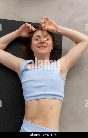 Une jeune femme se détache paisiblement sur un tapis, tenant une fleur pendant une séance de yoga Banque D'Images