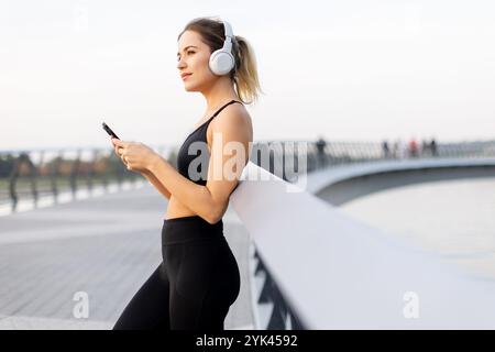 Une jeune femme se tient détendue sur un pont, écouteurs, savourant de la musique tout en se prélassant dans la douce lueur du coucher de soleil près du bord de l'eau. Banque D'Images