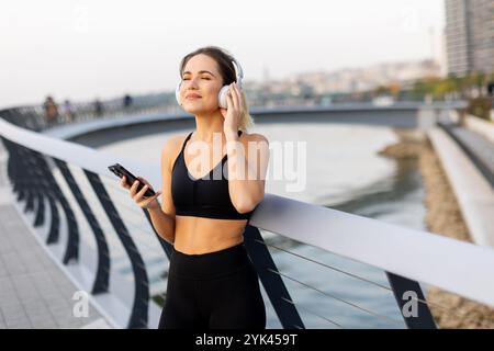 Une jeune femme se tient détendue sur un pont, écouteurs, savourant de la musique tout en se prélassant dans la douce lueur du coucher de soleil près du bord de l'eau. Banque D'Images