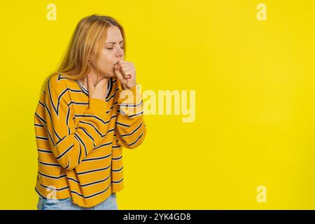 Femme blonde caucasienne malsaine en blouse toussant avec des symptômes de grippe saisonnière couvrant la bouche avec la main, se sentant malade, allergie, fièvre ou infection virale. Fille malade malheureuse isolée sur fond jaune Banque D'Images