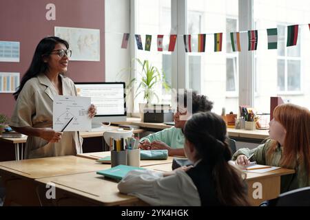 Jeune professeur confiant de grammaire anglaise parlant aux écoliers à la leçon à l'école primaire et expliquant les règles sur papier Banque D'Images