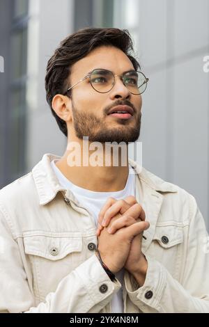 Homme indien priant les yeux fermés à Dieu demandant bénédiction, aide, pardon dehors. Un homme hindou arabe qui s'agrippait des mains souhaitant chance dans la rue du centre-ville. Styles de vie des gens d'affaires. Vertical Banque D'Images