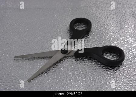 Ciseaux de bureau métalliques argentés avec oreilles noires sont situés sur un fond argenté texturé avec réflexion et points convexes de différentes formes. Banque D'Images