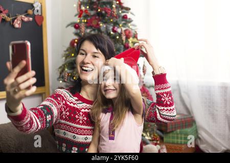 Maman et fille sont assis près de l'arbre de Noël, embrassant, prenant des photos et des selfies sur le téléphone, communiquant par communication vidéo, ayant f Banque D'Images