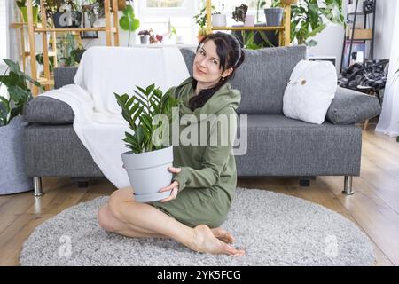 Zamioculcas sans prétention et populaire entre les mains d'une femme à l'intérieur d'une maison verte avec des collections de rayonnages de plantes domestiques. Récolte initiale Banque D'Images