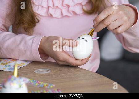 Une fille mignonne avec des oreilles de lapin rose fait un artisanat de Pâques, décore un œuf sous la forme d'une licorne avec des strass, de la corne, des fleurs à l'intérieur d'un Banque D'Images