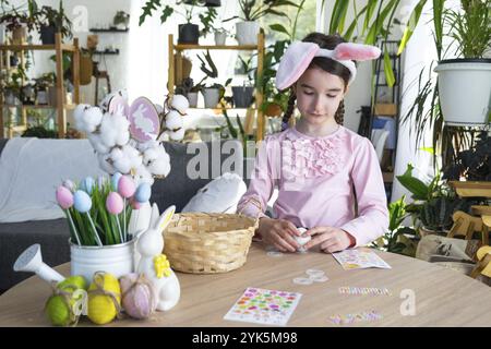 Une fille mignonne avec des oreilles de lapin rose fait un artisanat de Pâques, décore un œuf sous la forme d'une licorne avec des strass, de la corne, des fleurs à l'intérieur d'un Banque D'Images