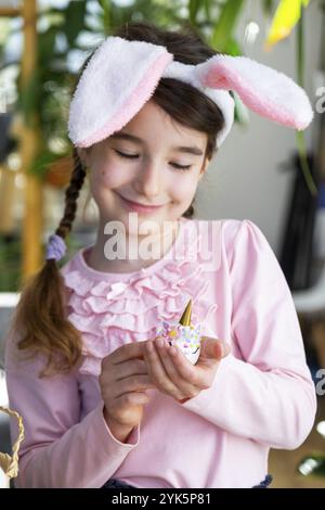 Une fille mignonne avec des oreilles de lapin rose fait un artisanat de Pâques, décore un œuf sous la forme d'une licorne avec des strass, de la corne, des fleurs à l'intérieur d'un Banque D'Images