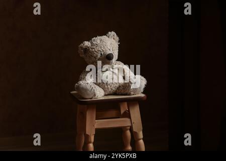 Ours en peluche solitaire assis sur une chaise. Un ours en peluche seul assis dans l'obscurité, vue de côté, jouet oublié Banque D'Images