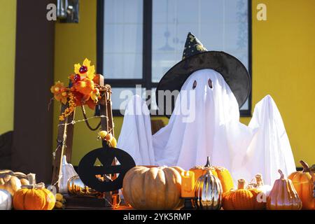 Enfant dans le drap de lit avec des fentes comme fantôme dans le chapeau de sorcière et le décor d'Halloween sur le porche de la maison à l'extérieur dans la cour de citrouille, lanterne, guirlande Banque D'Images