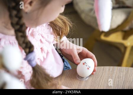 Une fille mignonne avec des oreilles de lapin rose fait un artisanat de Pâques, décore un œuf sous la forme d'une licorne avec des strass, de la corne, des fleurs à l'intérieur d'un Banque D'Images