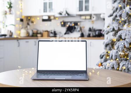 Ordinateur portable avec une maquette d'écran blanc dans la cuisine de Noël décorée blanche confortable avec des lumières de fée et un arbre de Noël. Télétravail saisonnier, interne Banque D'Images