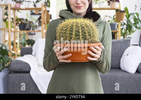 Grand échinocactus Gruzoni entre les mains d'une femme à l'intérieur d'une maison verte avec des collections de rayonnages de plantes domestiques. Production de cultures à domicile Banque D'Images