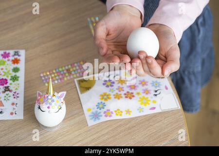 Une fille mignonne avec des oreilles de lapin rose fait un artisanat de Pâques, décore un œuf sous la forme d'une licorne avec des strass, de la corne, des fleurs à l'intérieur d'un Banque D'Images