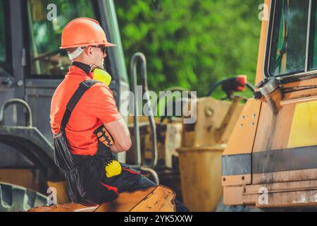 Un travailleur de la construction portant un casque et un équipement réfléchissant est assis sur une machine lourde pendant une pause sur un chantier de construction animé. Banque D'Images