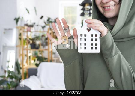 Une femme heureuse dans sa maison tient dans ses mains une figurine miniature d'une maison et la clé à l'intérieur. Projet de maison de rêve, achat immobilier, insura Banque D'Images