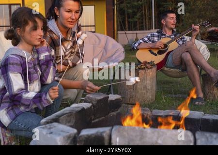 Une fille dans une chemise à carreaux rôtit des guimauves sur un feu dans la cour de la maison, père joue de la guitare. Soirée de réunion en famille près du feu de camp, Banque D'Images