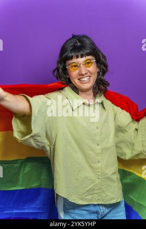 Portrait de studio avec fond violet d'une personne heureuse non binaire élevant le drapeau arc-en-ciel lgbt Banque D'Images