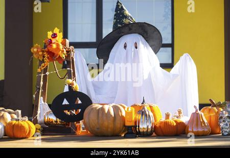Enfant dans le drap de lit avec des fentes comme fantôme dans le chapeau de sorcière et le décor d'Halloween sur le porche de la maison à l'extérieur dans la cour de citrouille, lanterne, guirlande Banque D'Images