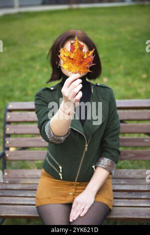 Une femme avec une feuille d'érable jaune tombé couvre son visage.Portrait d'automne sans visage sur un banc de parc.Ambiance d'automne Banque D'Images