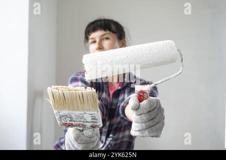 Femme perplexe dans le rouleau de peinture et la peinture blanche pour les murs dans les mains gros plan portrait. Travaux de construction et réparations cosmétiques dans la maison, peinture murale, TI Banque D'Images