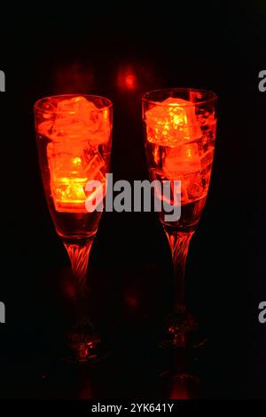 Des glaçons rouges brillants flottent dans des verres à champagne avec une boisson. Les objets en plastique fabriqués sous forme de glaçons avec une diode intégrée s'illuminent. Banque D'Images