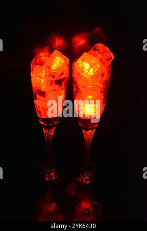 Des glaçons rouges brillants flottent dans des verres à champagne avec une boisson. Les objets en plastique fabriqués sous forme de glaçons avec une diode intégrée s'illuminent. Banque D'Images