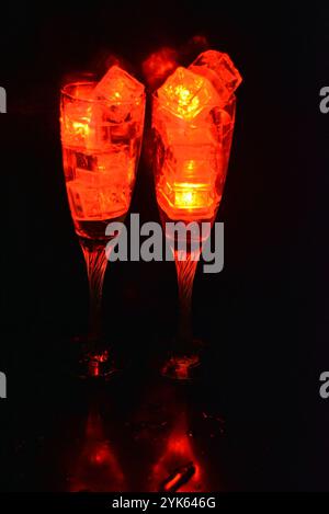 Des glaçons rouges brillants flottent dans des verres à champagne avec une boisson. Les objets en plastique fabriqués sous forme de glaçons avec une diode intégrée s'illuminent. Banque D'Images