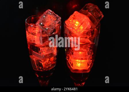 Des glaçons rouges brillants flottent dans des verres à champagne avec une boisson. Les objets en plastique fabriqués sous forme de glaçons avec une diode intégrée s'illuminent. Banque D'Images