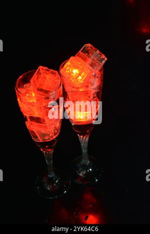 Des glaçons rouges brillants flottent dans des verres à champagne avec une boisson. Les objets en plastique fabriqués sous forme de glaçons avec une diode intégrée s'illuminent. Banque D'Images