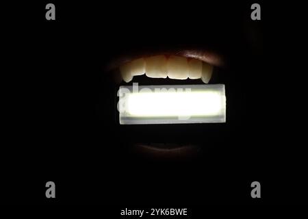 La jeune fille tient une petite lampe de poche dans ses dents dans l'obscurité complète, brille une lampe de poche, sans lumière Banque D'Images